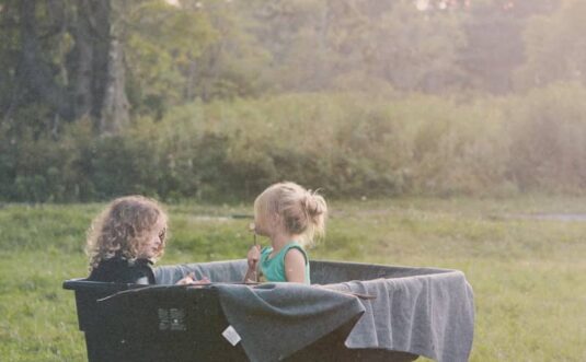 Two children sit in a wheelbarrow outdoors.
